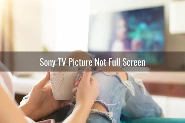 Woman in jeans sitting on sofa and holding a coffee mug watching TV