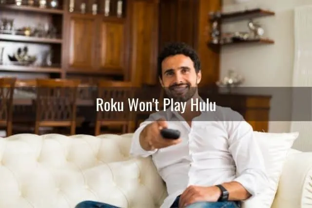 Male sitting on white sofa with TV remote in hand pointing at TV